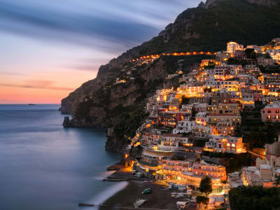 Positano village glowing with warm lights at dusk reflected in calm Mediterranean sea Amalfi Coast Italy