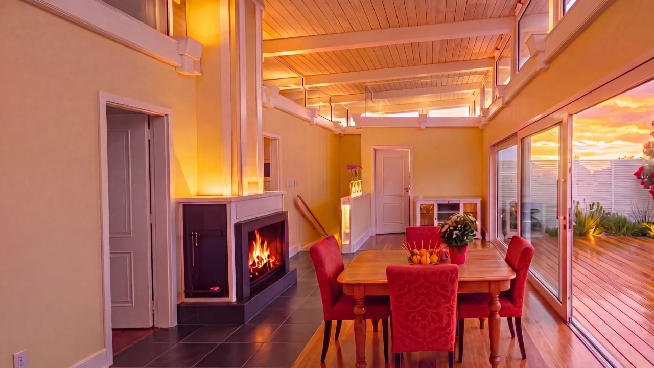 Annex dining — fireplace blazing, red chairs at timber table, sliding doors to sunset deck, T&G ceiling, Hawke's Bay NZ