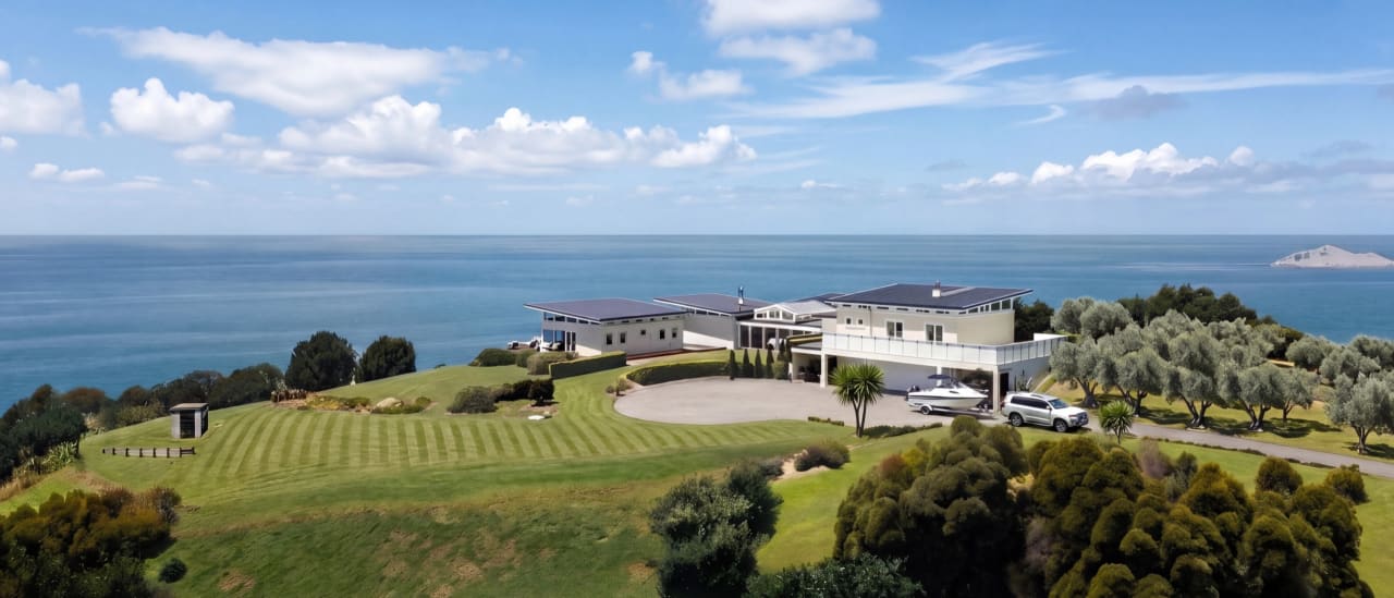 Panoramic aerial of Bay House showing both wings, solar roof, manicured lawn, carport with boat, and Pacific ocean to horizon
