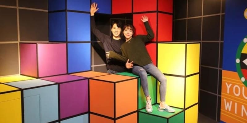Stacks Of Colorful Cube Boxes With A Couple Seated On One, Waving And Smiling.