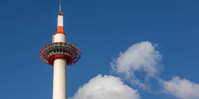 A Tall Tower With White And Red Coloring Has A White Antenna On Top. It Stands Against A Blue Sky With Clouds.