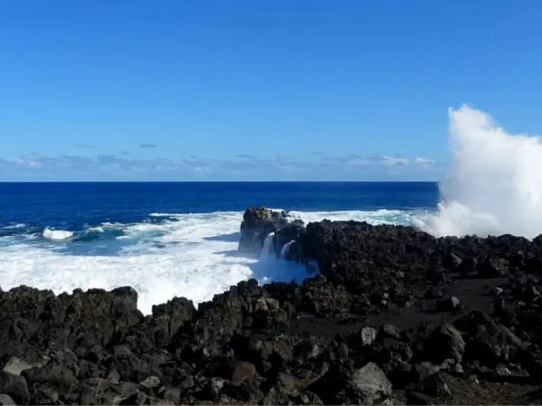 Gerbe d'écume blanche jaillissant entre des blocs de basalte noir, océan bleu sous un ciel dégagé