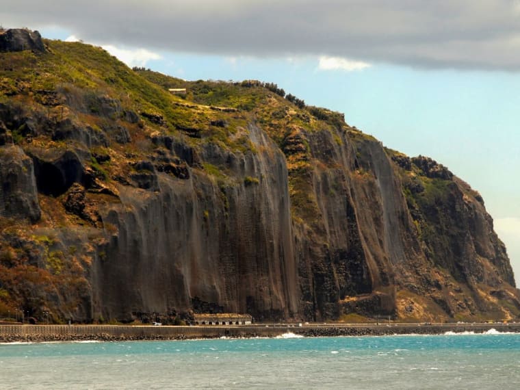 Haute falaise brune plongeant dans l'océan, avec une route qui longe la mer à son pied