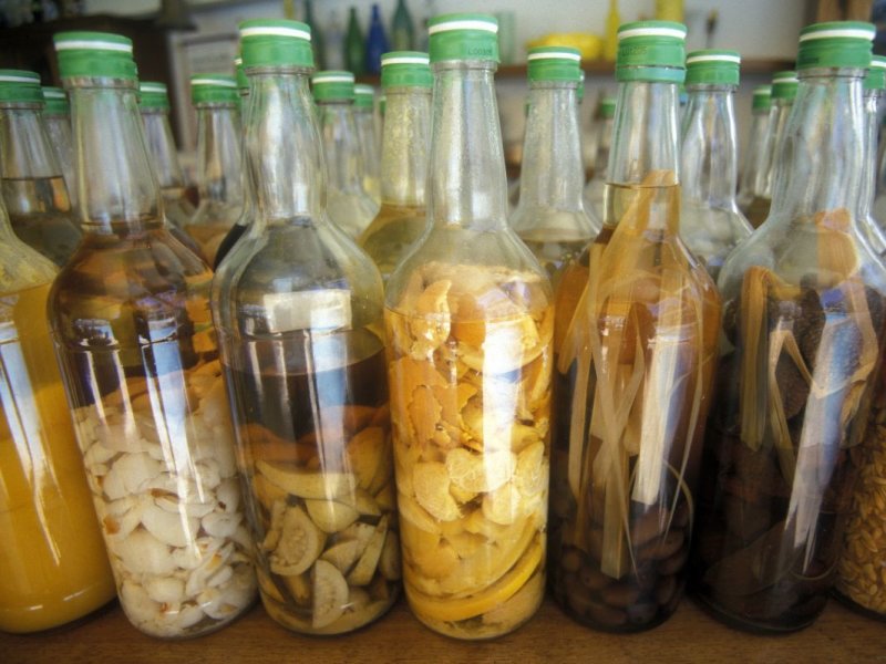 Rangée de bouteilles bouchées de vert où macèrent des tranches d'agrumes, des bâtons, des graines et des feuilles dans du rhum