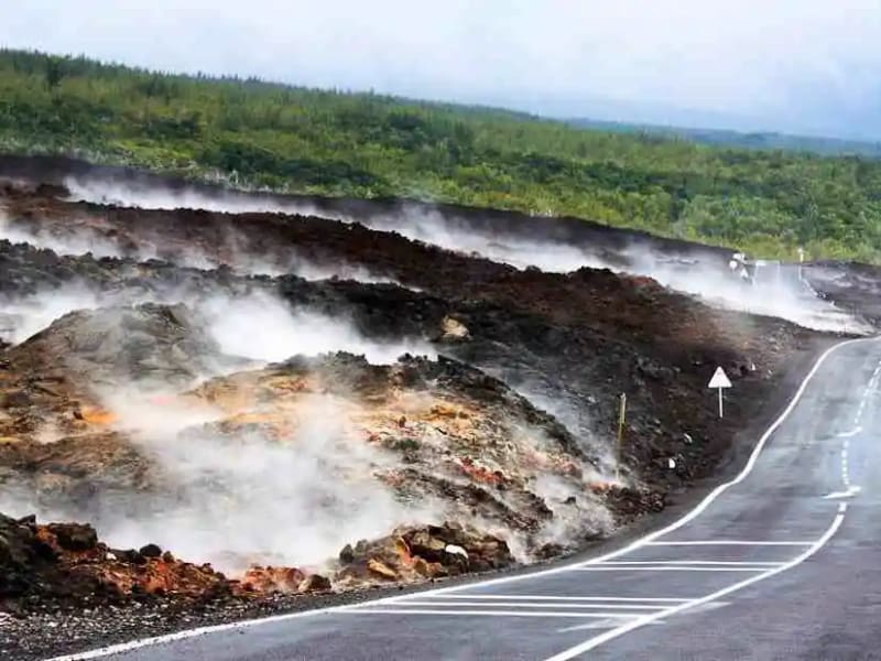 Route goudronnée longeant une coulée de lave qui fume encore, panneau de signalisation et forêt verte au loin