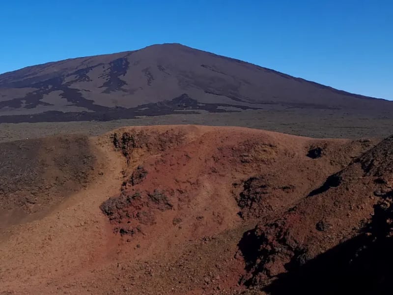 Cône de scories rouge au premier plan et grande pente sombre du Piton de la Fournaise sous un ciel bleu sans nuage