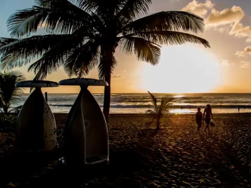 Deux silhouettes marchant sur une plage de sable sombre au coucher du soleil, palmiers et coques de pirogues dressées au premier plan