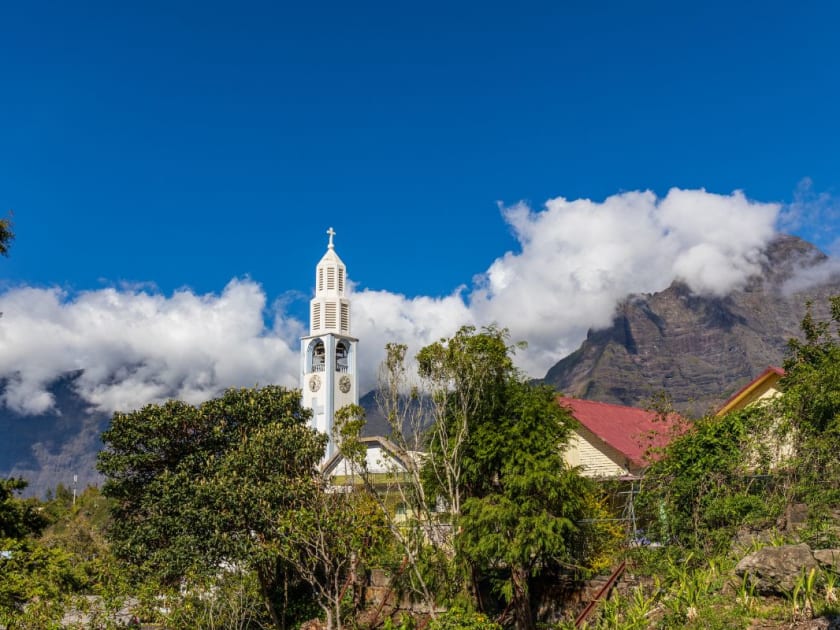 Clocher blanc d'une église dépassant de la végétation, toits rouges et crêtes à demi prises dans les nuages