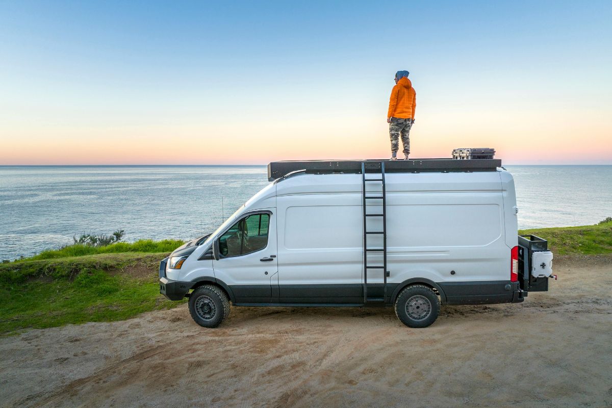 Voyageur sur le toit d'un van aménagé Ford Transit blanc face à l'océan Indien au coucher de soleil, road-trip à La Réunion