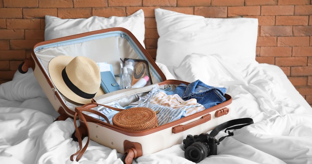 Valise rigide ouverte sur un lit, contenant un chapeau de paille, des vêtements et accessoires pour un voyage tropical