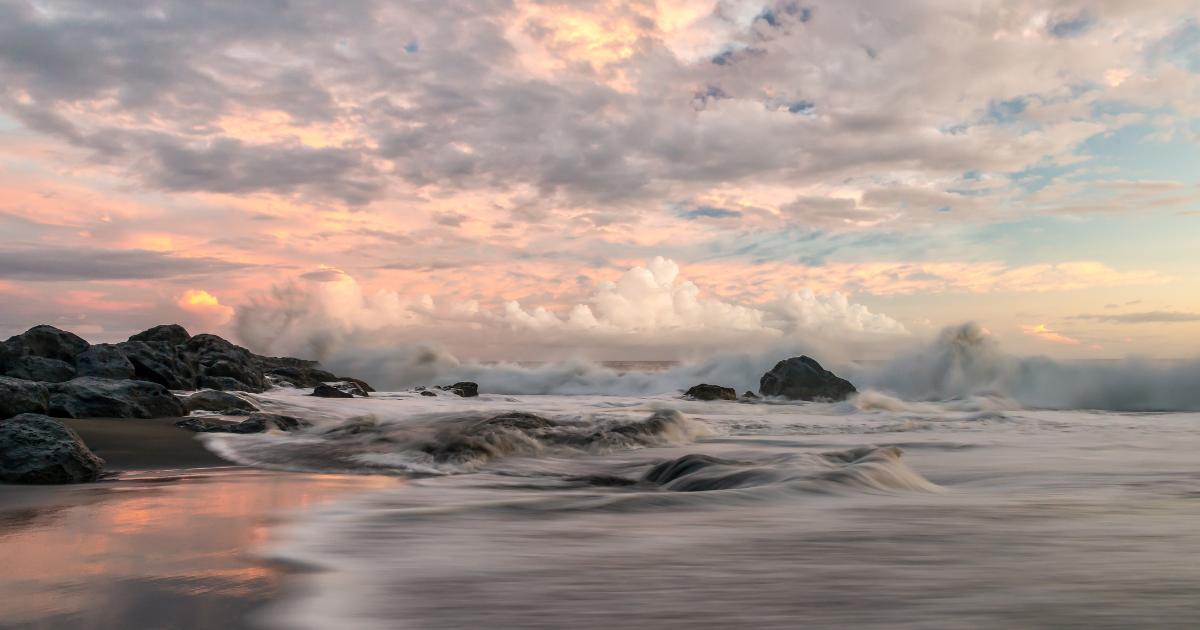 Plage de sable noir de La Réunion en février au coucher du soleil, vagues et ciel rose chargé