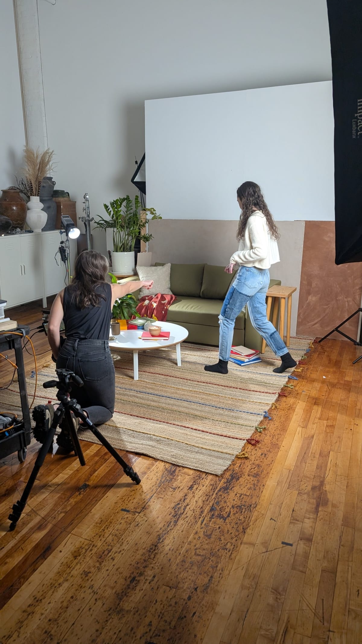 A photographer crouched behind a tripod as a model walks into a lit living room set