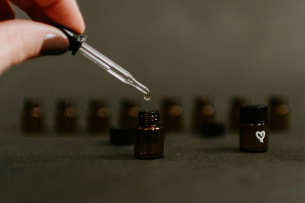 A hand using a glass pipette dropper above a small amber fragrance bottle, a single drop falling, rows of more amber bottles softly out of focus in the background