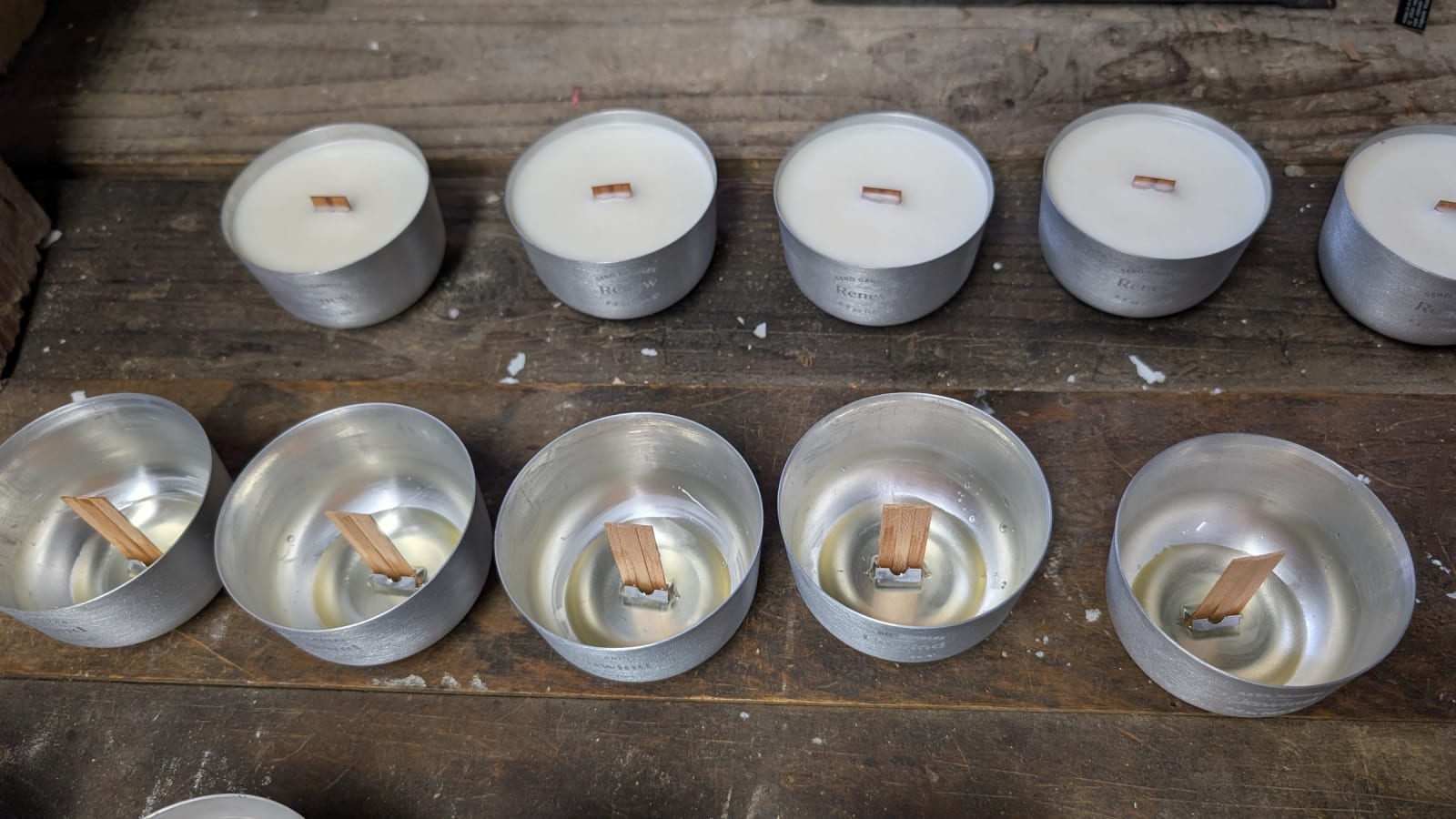 Two rows of Sero candle inserts on a workshop bench. Top row poured and curing; bottom row empty, showing welded aluminum wick clips at the base of each cup with wood wicks standing upright.