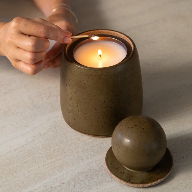 A hand lighting the Highbrow Hippie hand-thrown ceramic refillable candle, its domed lid resting beside it.
