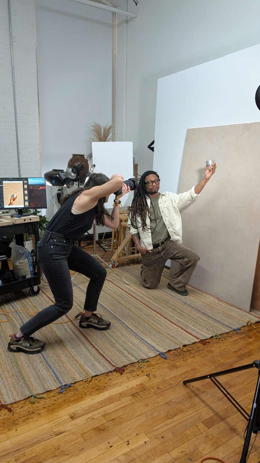 A photographer bent low with a camera, shooting a model who is holding a candle against a plaster-finished board on a stand