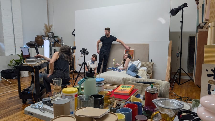 A bed set built on a wooden pallet frame in a studio, surrounded in the foreground by prop buckets, mugs and paint cans