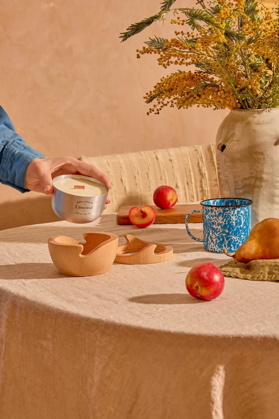 A hand lowering an Unwind aluminum Sero candle onto a linen-covered table set with fruit, an enamel mug and dried mimosa