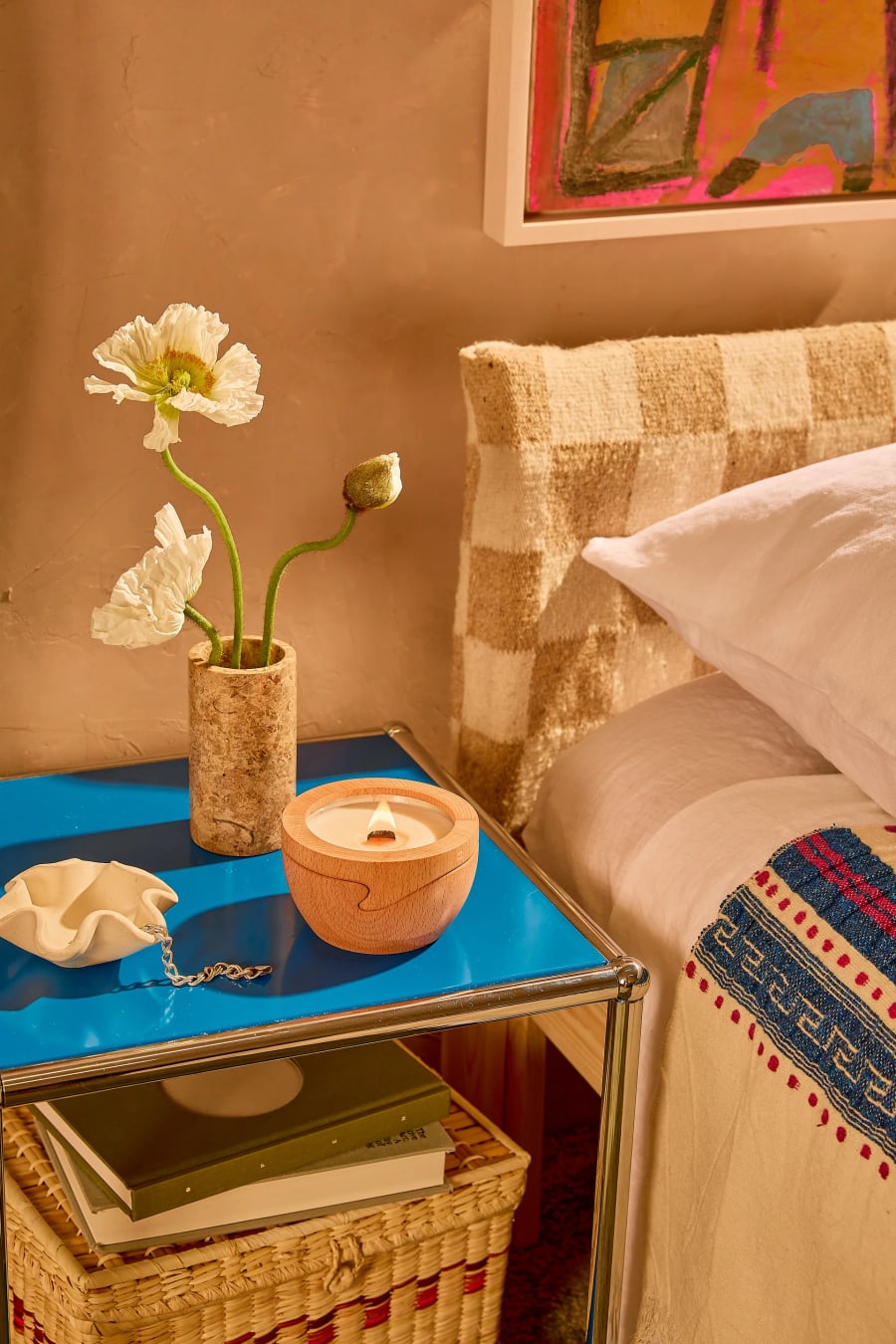 A lit Sero candle in its beech container on a blue nightstand beside white poppies, next to a made bed