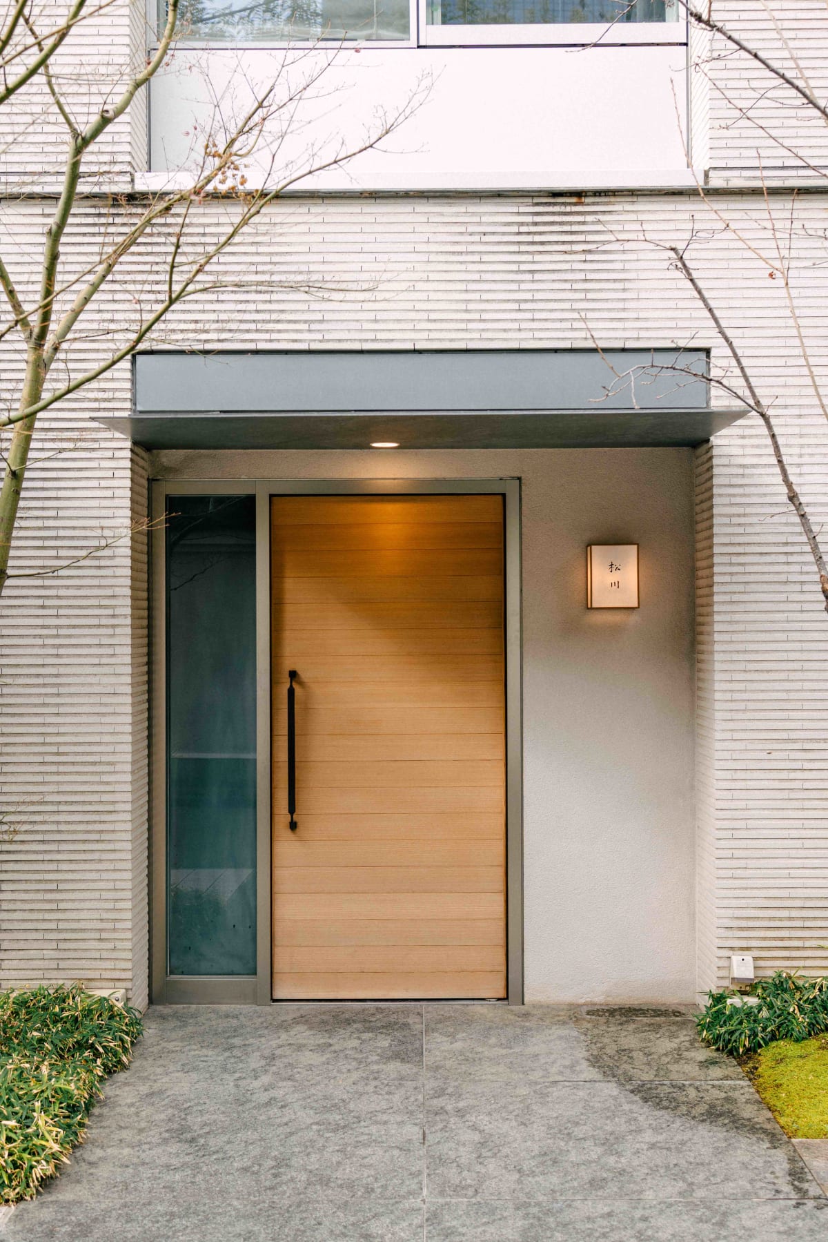 Matsukawa, Tokyo: the entrance, a pale wooden door beside a small lantern sign, bare trees on either side