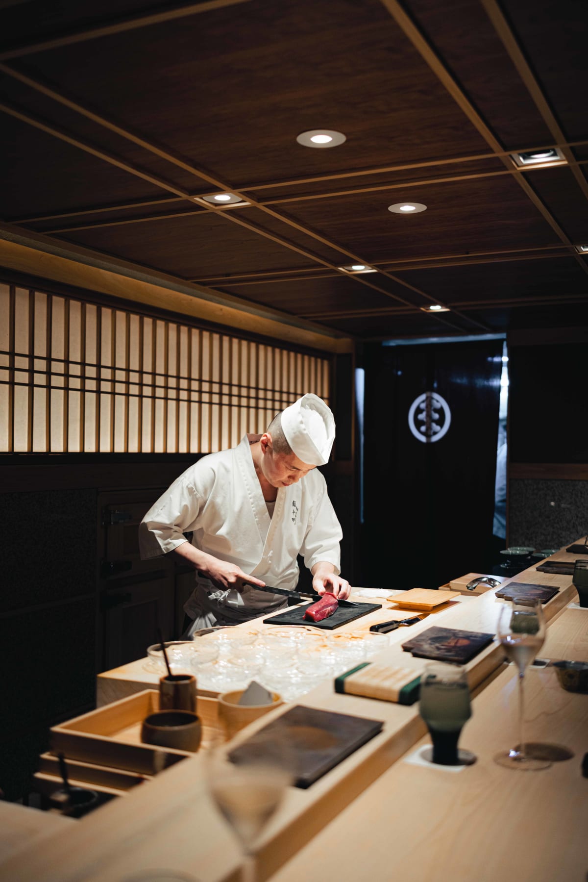 Sushi Juyon by Saito, Seoul: the long room on the second visit, the counter stretching beneath the shoji windows and the crested noren, the taisho slicing tuna and plating at its centre