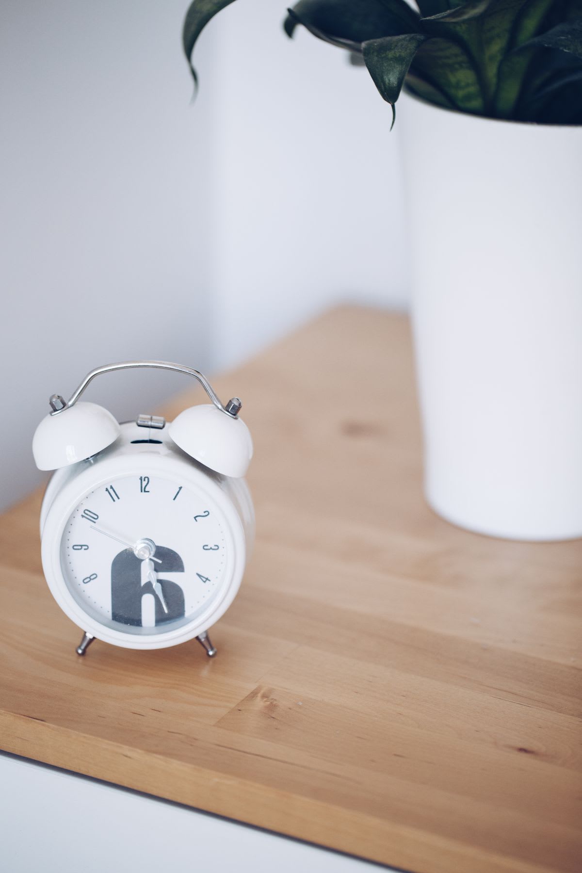 A small white alarm clock on a wooden table beside a potted plant