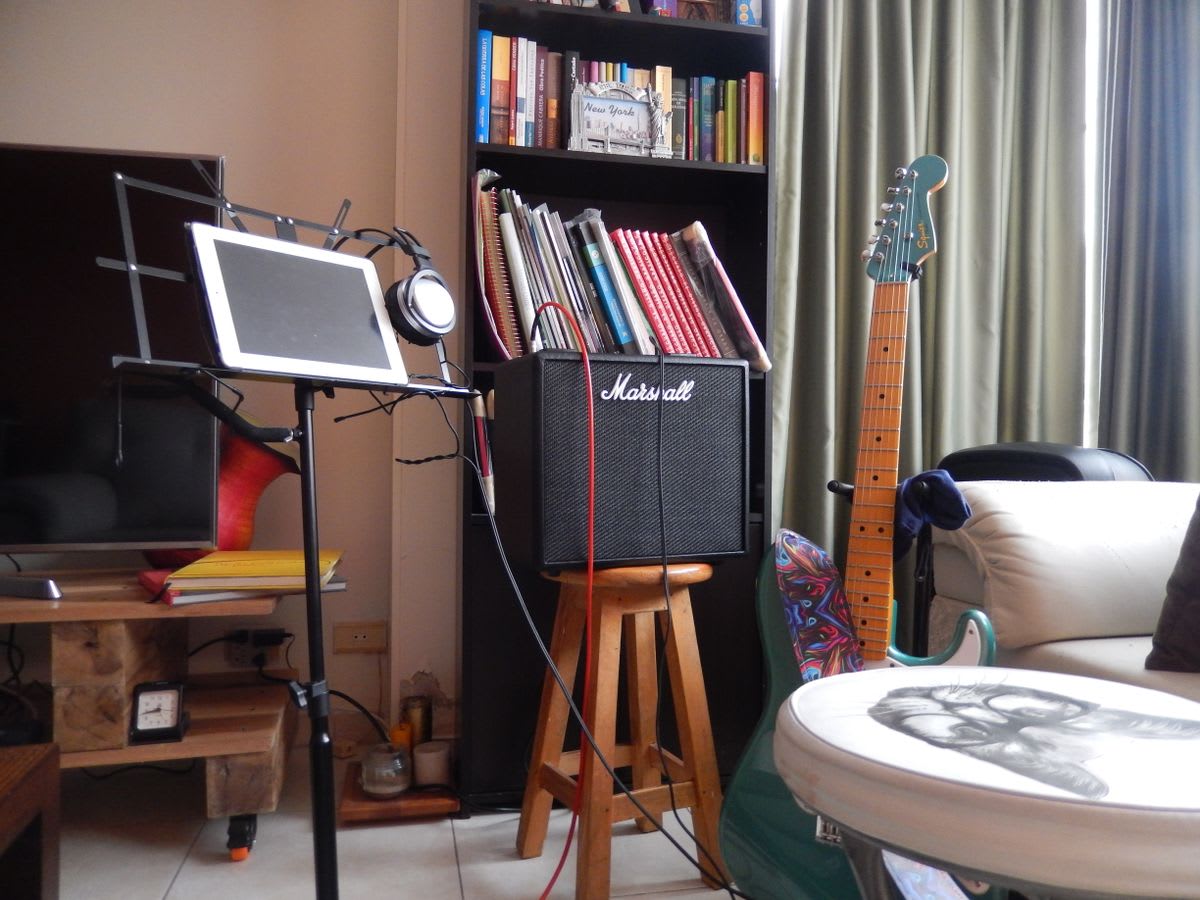 A tablet on a music stand beside a guitar and amplifier in a home rehearsal setup