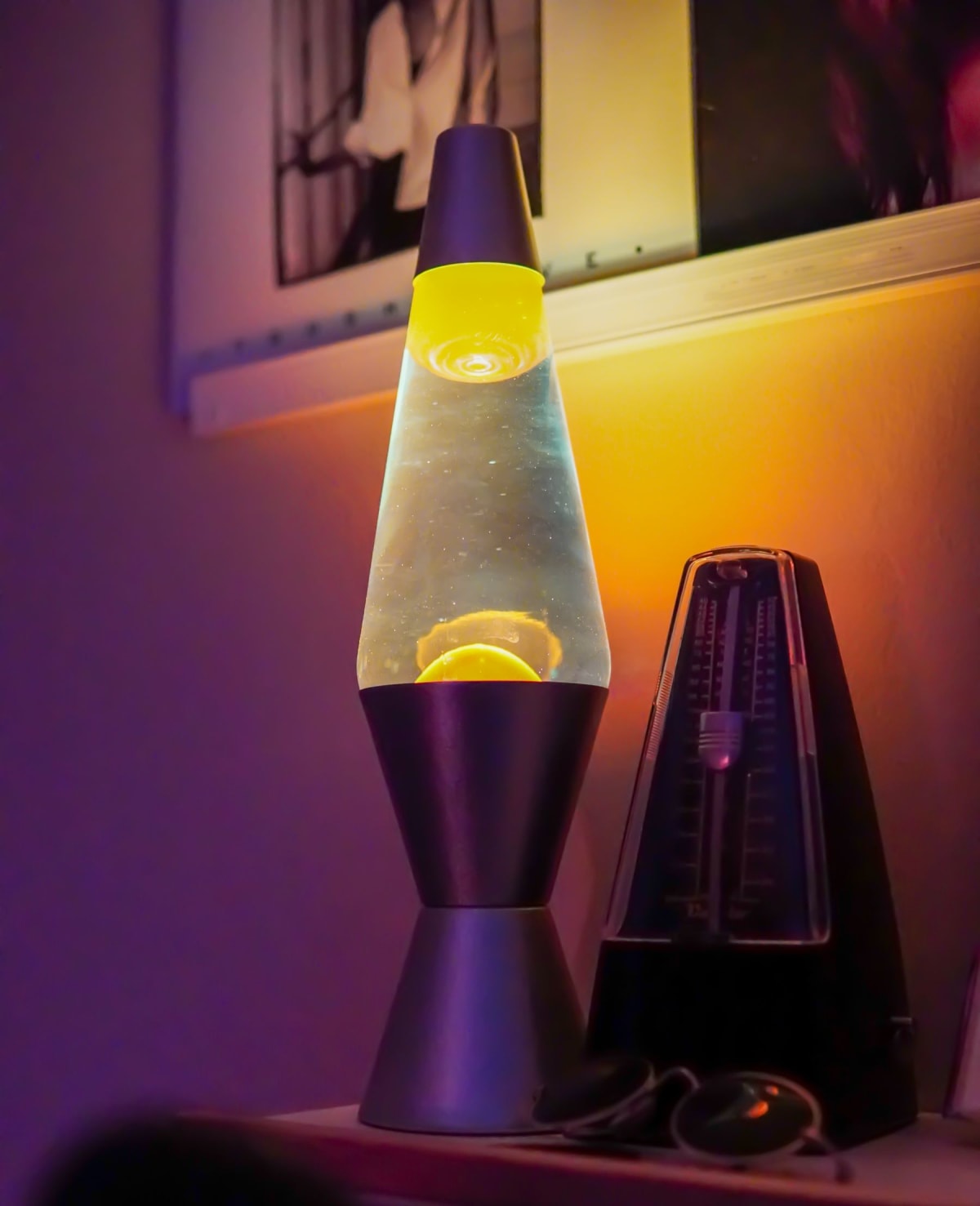 Black mechanical pendulum metronome beside a glowing lava lamp in a home music space