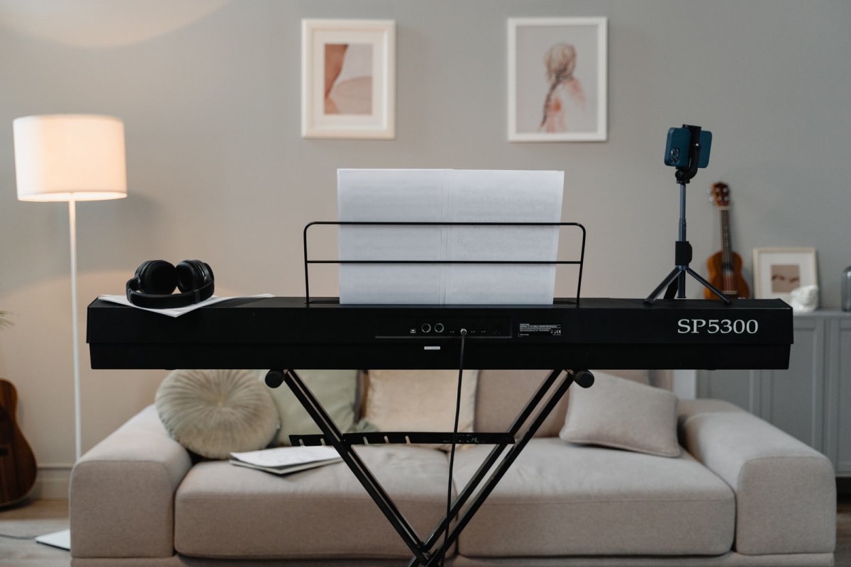 A digital piano on a stand with headphones, sheet music and a phone tripod in a living room