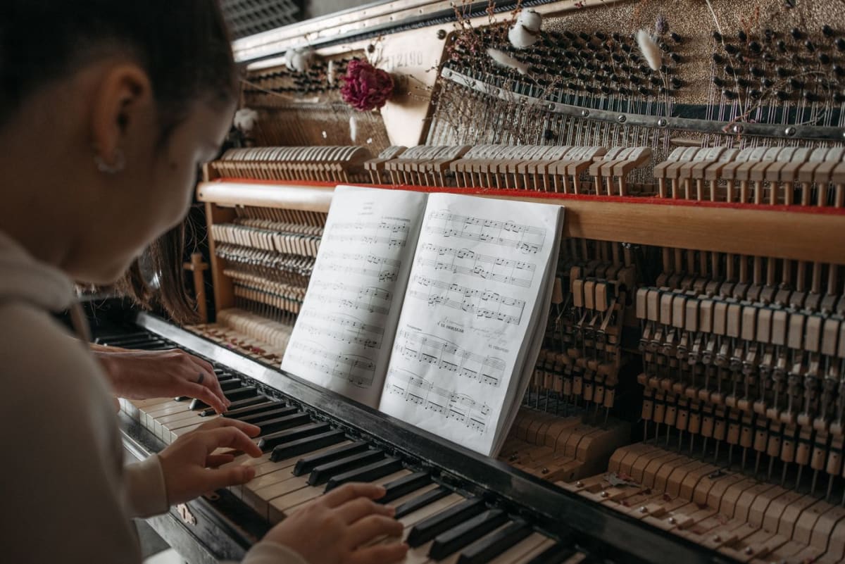 A pianist reading an open music book while playing alongside another person at an upright piano