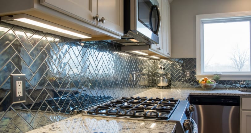 Modern kitchen with stainless appliances, granite countertop, and glossy herringbone tile backsplash under cabinet lighting