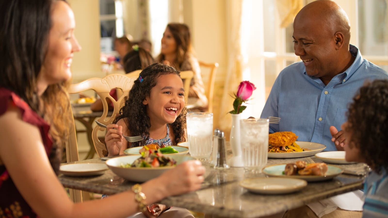 Family laughing over lunch at Grand Floridian Cafe in Disney's Grand Floridian Resort & Spa, with salads, a breaded cutlet and a pink rose