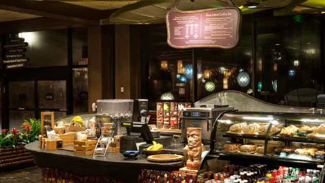 Kona Island coffee bar at Disney's Polynesian Village Resort with a pastry case, a carved tiki figure on the counter and a lit menu board above