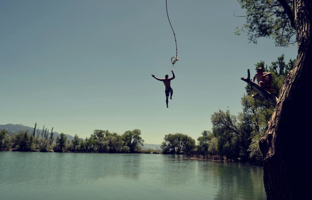 Man jumps on a rope into a blue lake, Travel Buddy is waiting on a branch.