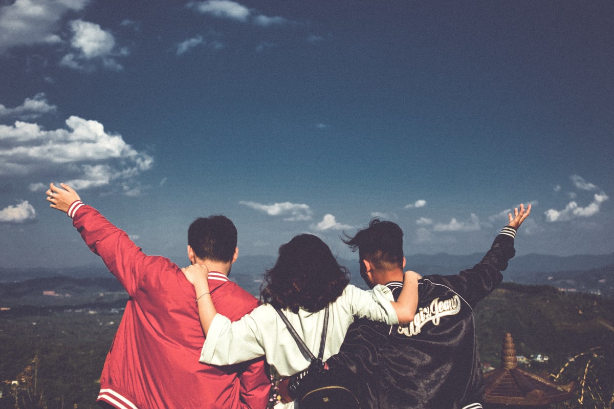 three friends standing on top of a mountain looking down, woman has her arms around her two male friends 