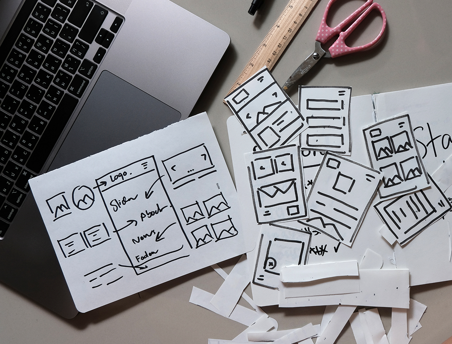 Hand drawn layout sketches cut out and scattered across a desk beside a laptop, ruler and scissors