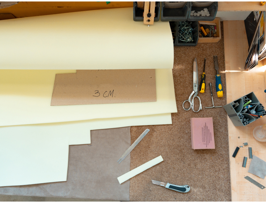 Foam and cardboard sheets part cut on a workbench beside scissors, a craft knife, a metal rule and a template marked 3 CM