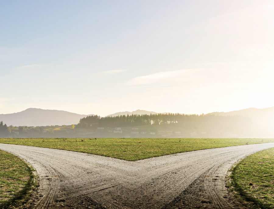 A gravel track splitting into two paths across a green field at sunrise, with hills and mist in the distance