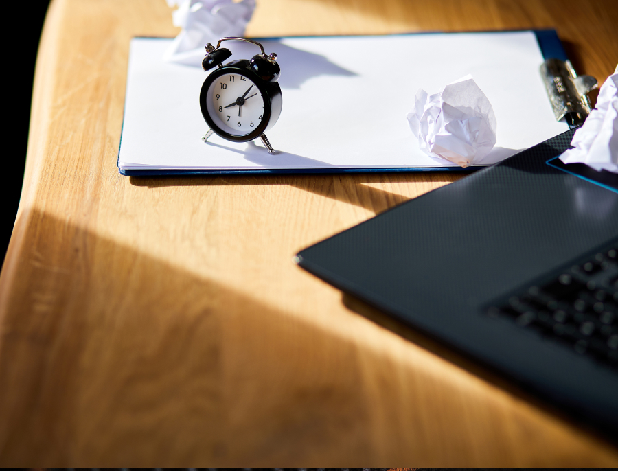 A small alarm clock standing on a blank notepad on a wooden desk, beside crumpled paper and the corner of a laptop in hard morning light