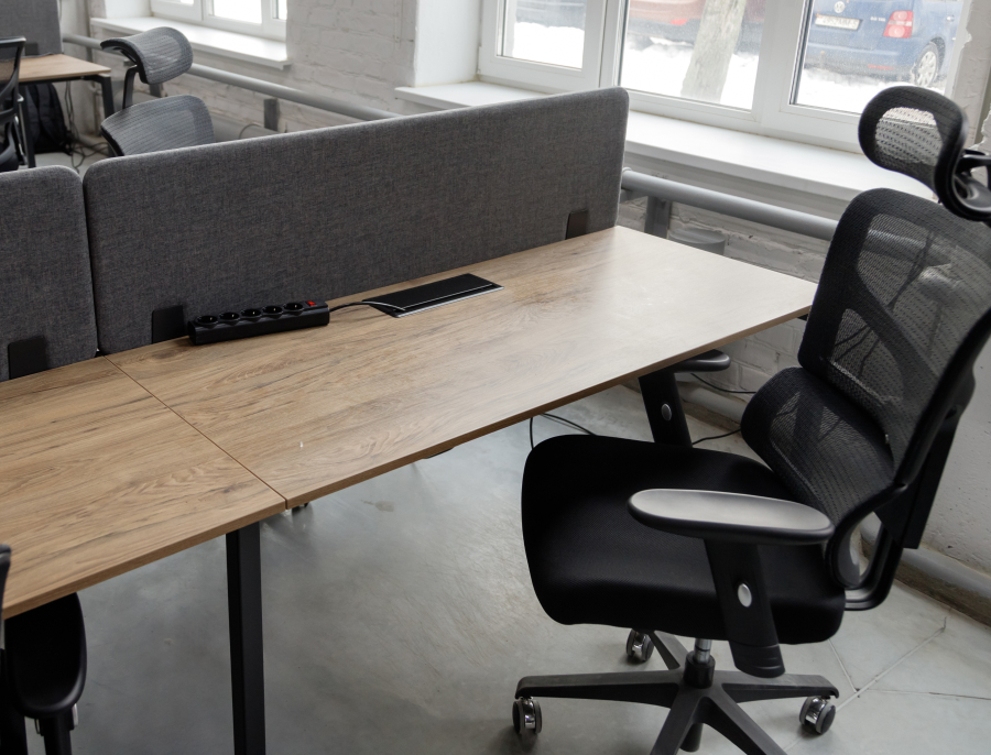 An empty office chair at a bare shared desk holding only a power strip, beside a window