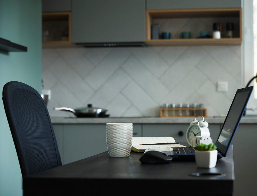 A laptop, notebook, mug and small alarm clock set up on a kitchen counter with a desk chair pulled up to it, the kitchen visible behind