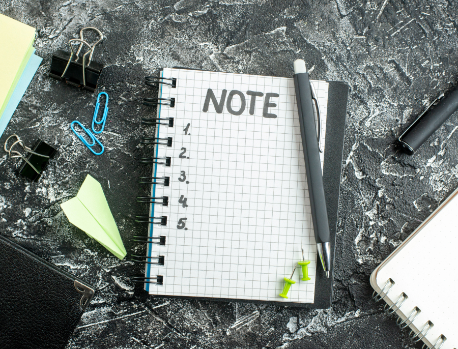 An overhead view of a spiral notepad headed NOTE with an empty numbered list, surrounded by pens, paper clips and pins