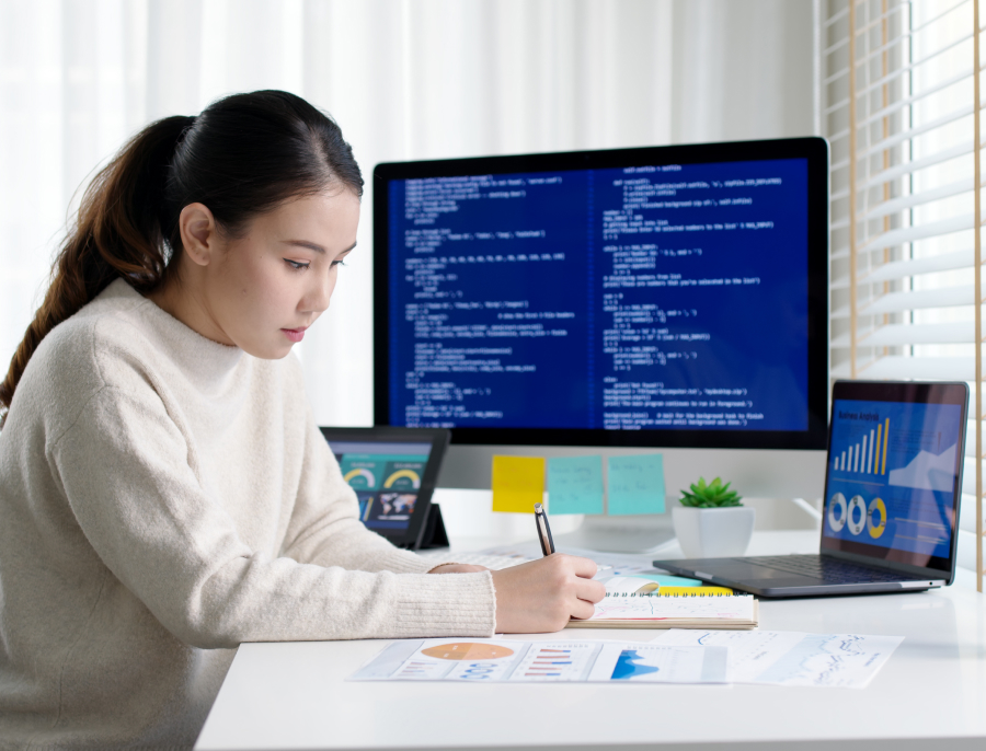 A woman writing in a notebook at a desk with a large monitor, a laptop and a tablet all showing different screens