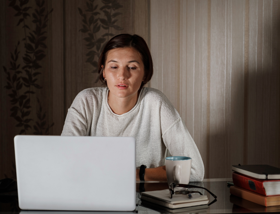 A woman watching her laptop in a dimly lit room in the evening, a cup and a stack of books beside her