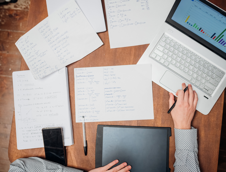 An overhead view of handwritten pages spread across a wooden desk beside a laptop and a graphics tablet