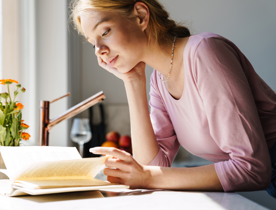 A woman resting her chin on her hand while reading an open book by a bright window