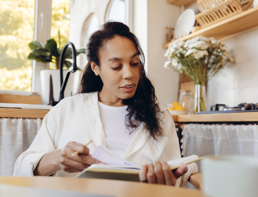 A woman writing in an open notebook at a kitchen table, morning light through the window behind her