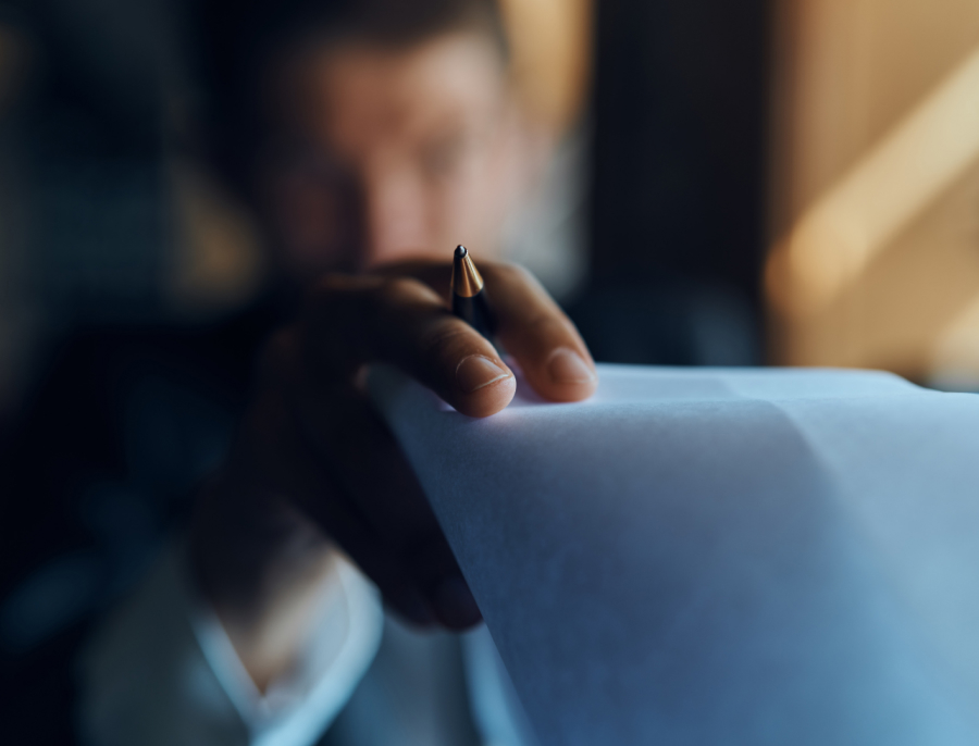 A person holding a pen above a sheet of paper, seen close up with the face out of focus behind it