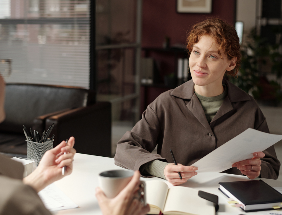 Two people talking across a desk with documents and notebooks between them, one listening while the other speaks