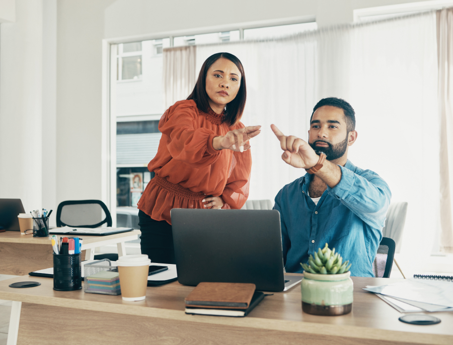 Two colleagues at a desk in an office, one standing and one seated, both gesturing while talking